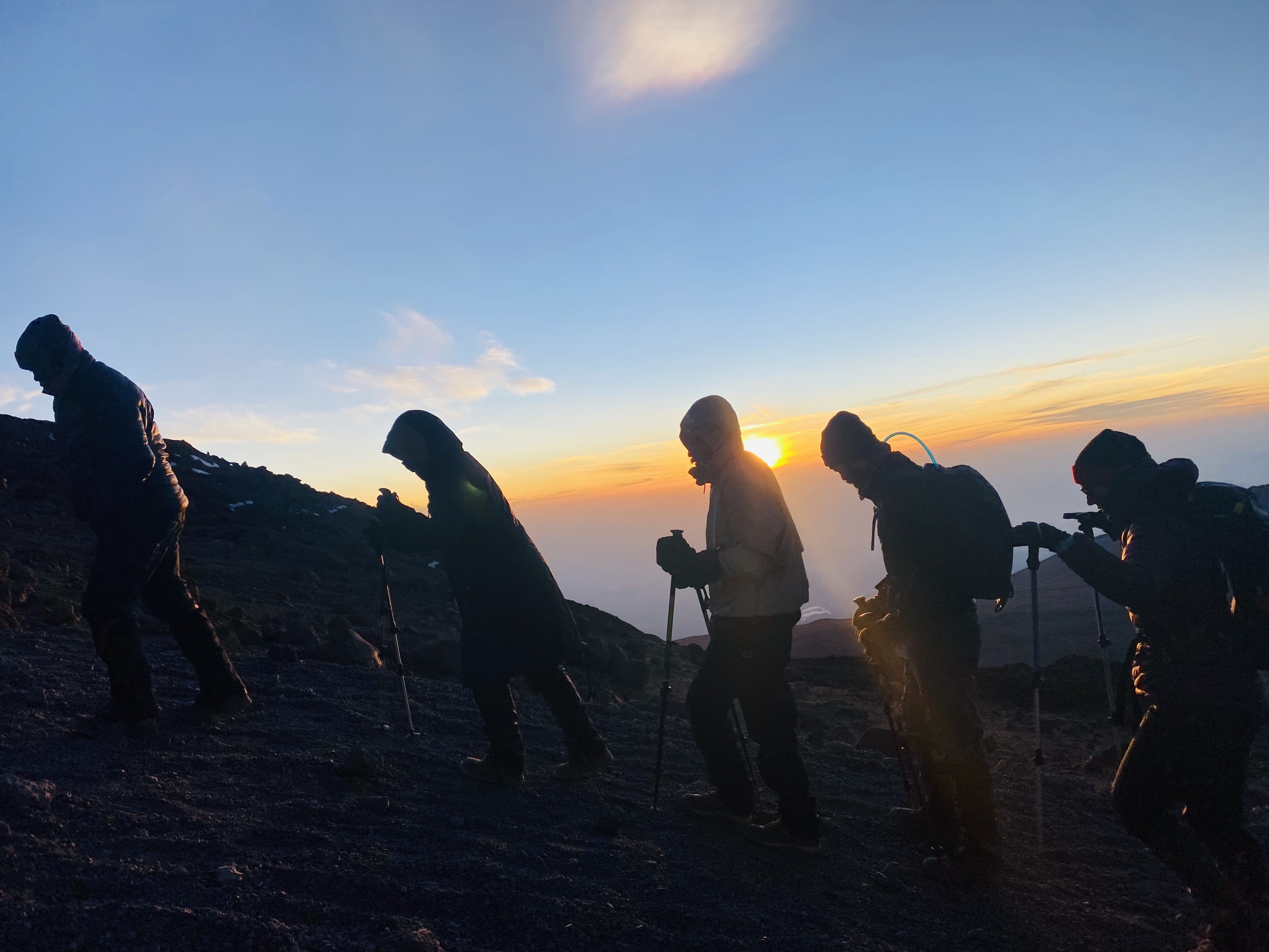 Silhouetted climbers with trekking poles ascending in the pre-dawn light of a summit sunrise
